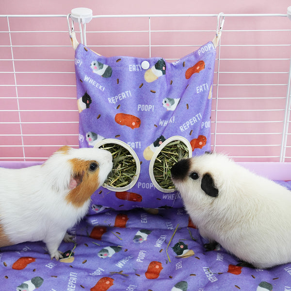 deux cochons d'inde mangeant du foin dans un sac à foin violet aux motifs caca de la marque Kavee dans une cage sur un tapis polaire